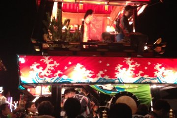 The robed figure atop the "dashi" is the neighborhood god. People on the lower level play taiko drums and flutes
