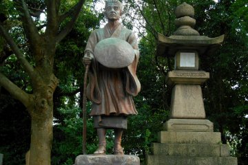 <p>Statue of Matsuo Basho, a famous poet in the Edo Period, standing in the corner of the shrine grounds with a stone lantern and tall trees</p>