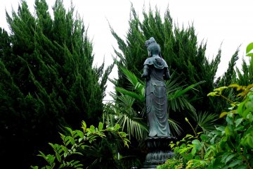 <p>A statue of Kannon peeps between overgrown cypress trees</p>