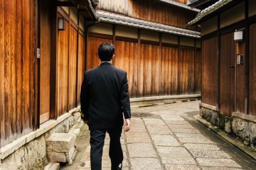 <p>Man in a modern suit walks through the backdrop of traditional houses.&nbsp;</p>