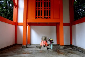 Patung Ojizo tampak sangat kecil dikeliling bangunan merah dan putih disekitarnya 