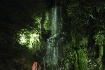 <p>An orange statue of&nbsp;Fudō-Myō-ō (immovable&nbsp;wisdom king) standing steadfast in front of the illuminated green waterfall.</p>