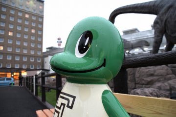 <p>Jurassic character mascot 'Rapt' sitting on a 'Jurassic Bench' placed in a open space in front of JR Fukui Station</p>