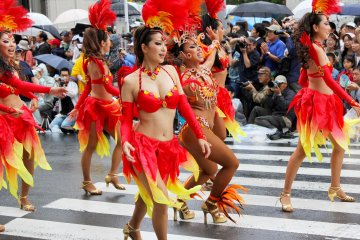 The dancers have various color coordinated outfits with one samba outfit dancer surrounded by costumed dancers.&nbsp;