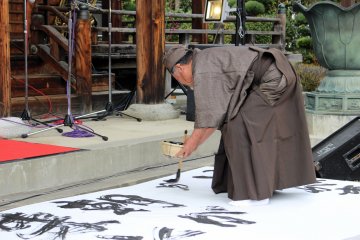 <p>The head of the Imai-cho Neighborhood Association (the organizer of the event) writes out a prayer and message in the welcoming calligraphy performance while a jazz band from the medical university plays a walking number</p>