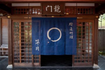 The restaurant entrance lies behind a blue noren curtain nestled behind some trees