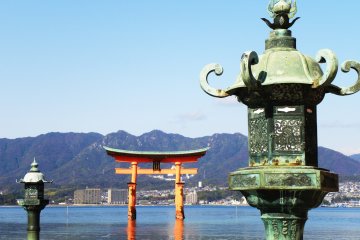 Una linterna de bronze dai-toro con las montañas y el Tori Flotante en el fondo. La linterna en la izquierda se encuentra en realidad entre otras dos linternas. ¿Notaste la diferencia en los ornamentos metálicos sobre cada linterna?