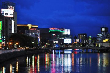 CIty lights mirrored by the river below creating a stream of lights. This area, Nakasu, borders Tenjin.